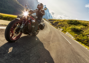 A man wearing a helmet and leather jacket riding a motorcycle down a scenic road, symbolizing a dedicated motorcycle owner.